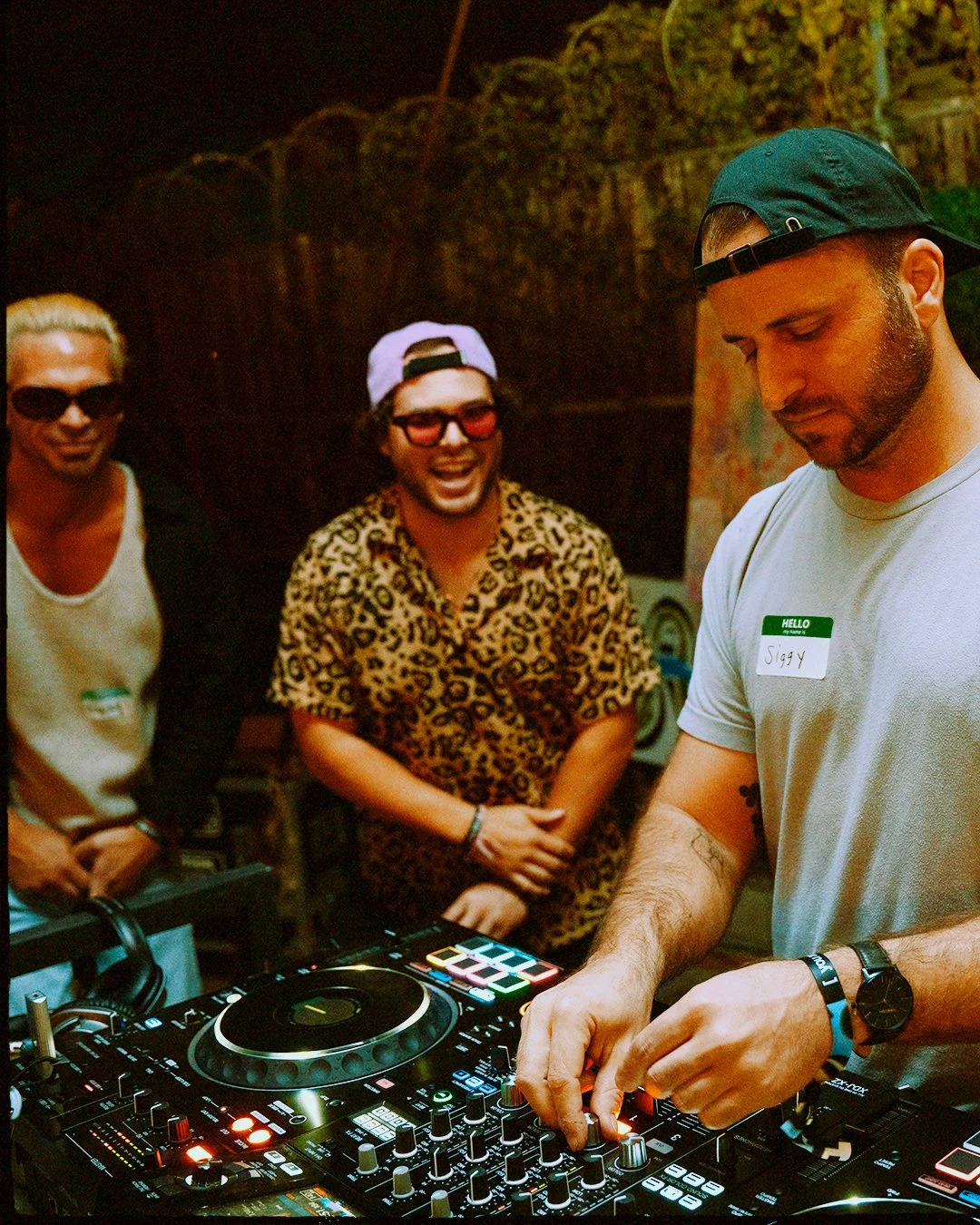 A DJ with a beard and baseball cap operates a DJ mixer at a party, with three people in the background, two men smiling and one woman in sunglasses and a leopard print shirt.