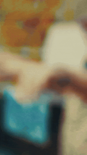 Blurred image showing a person in a blue shirt, possibly holding an object indoors with some colorful background elements.