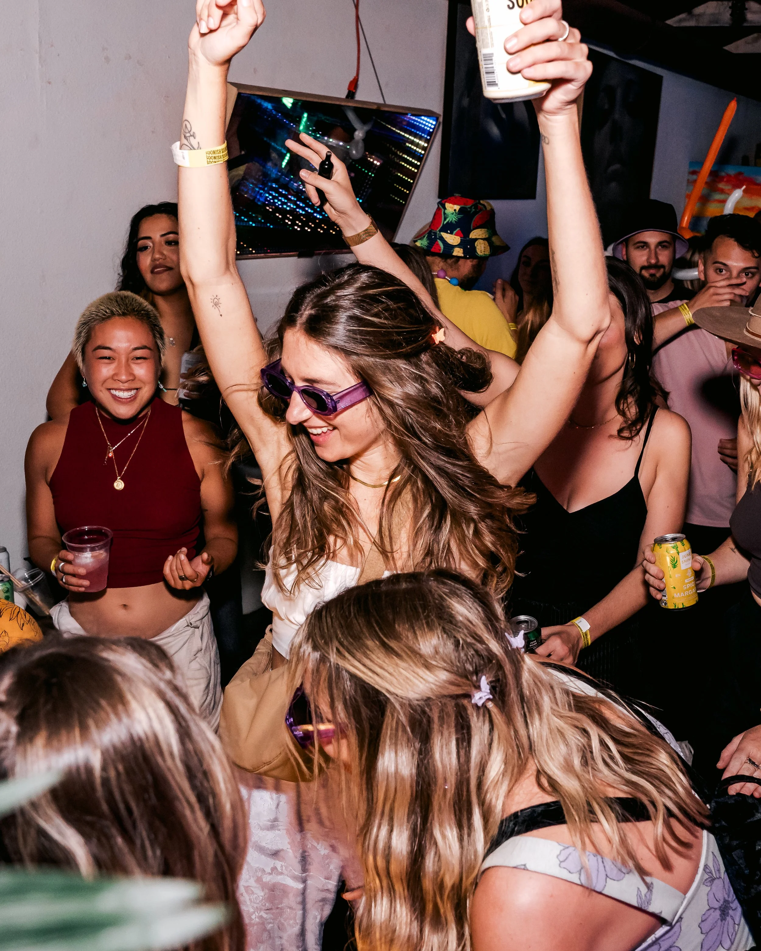 People dancing and celebrating at a party, with one woman in sunglasses raising her hands and smiling.