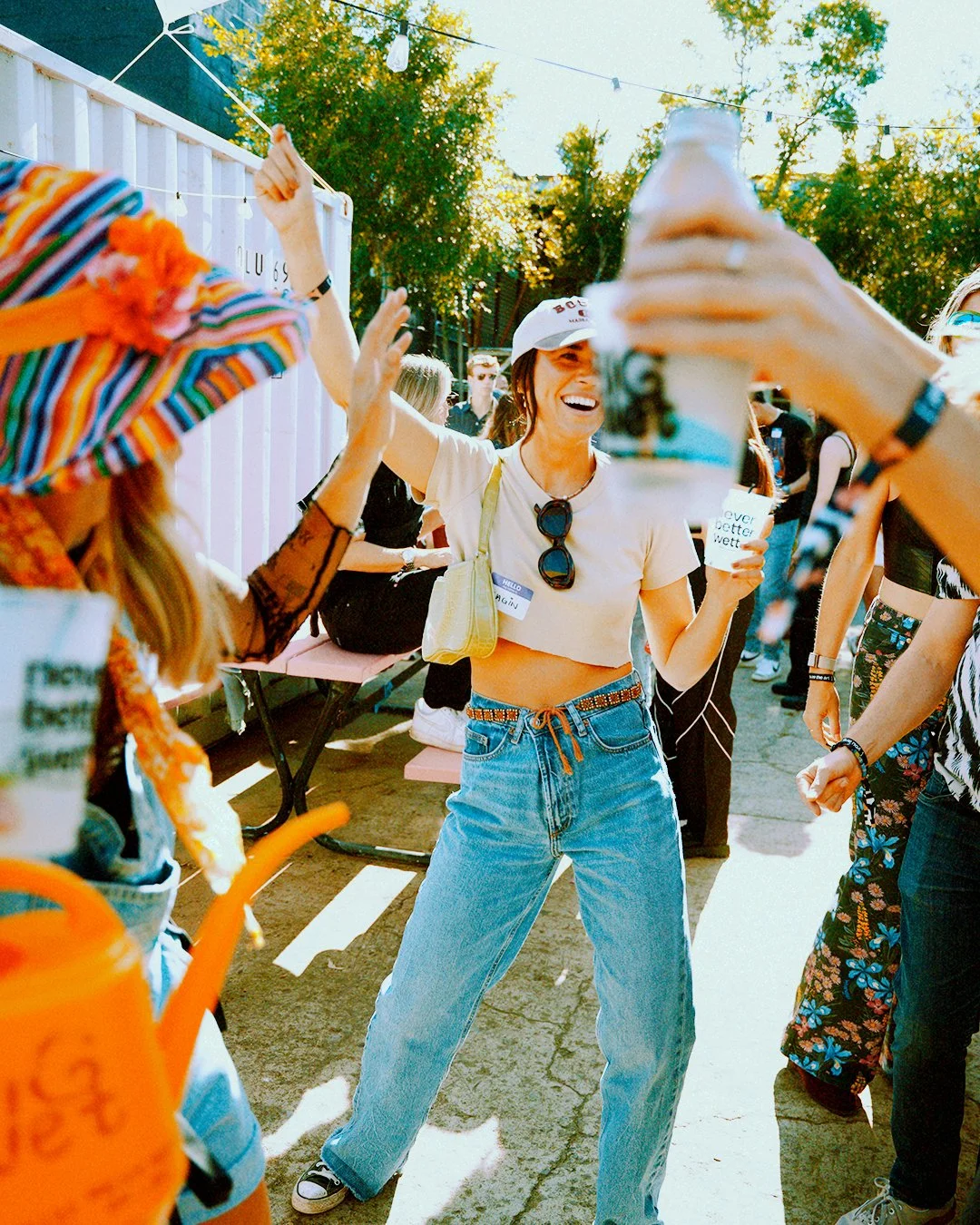 People celebrating outdoors at a festival or gathering, high-fiving and laughing on a sunny day with trees and string lights overhead.