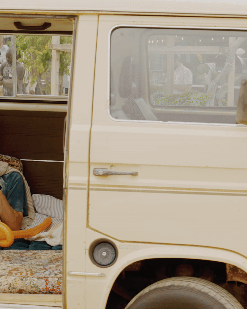 Partial view of a vintage beige van, showing the front passenger door and window, with some people visible through the windows outside.