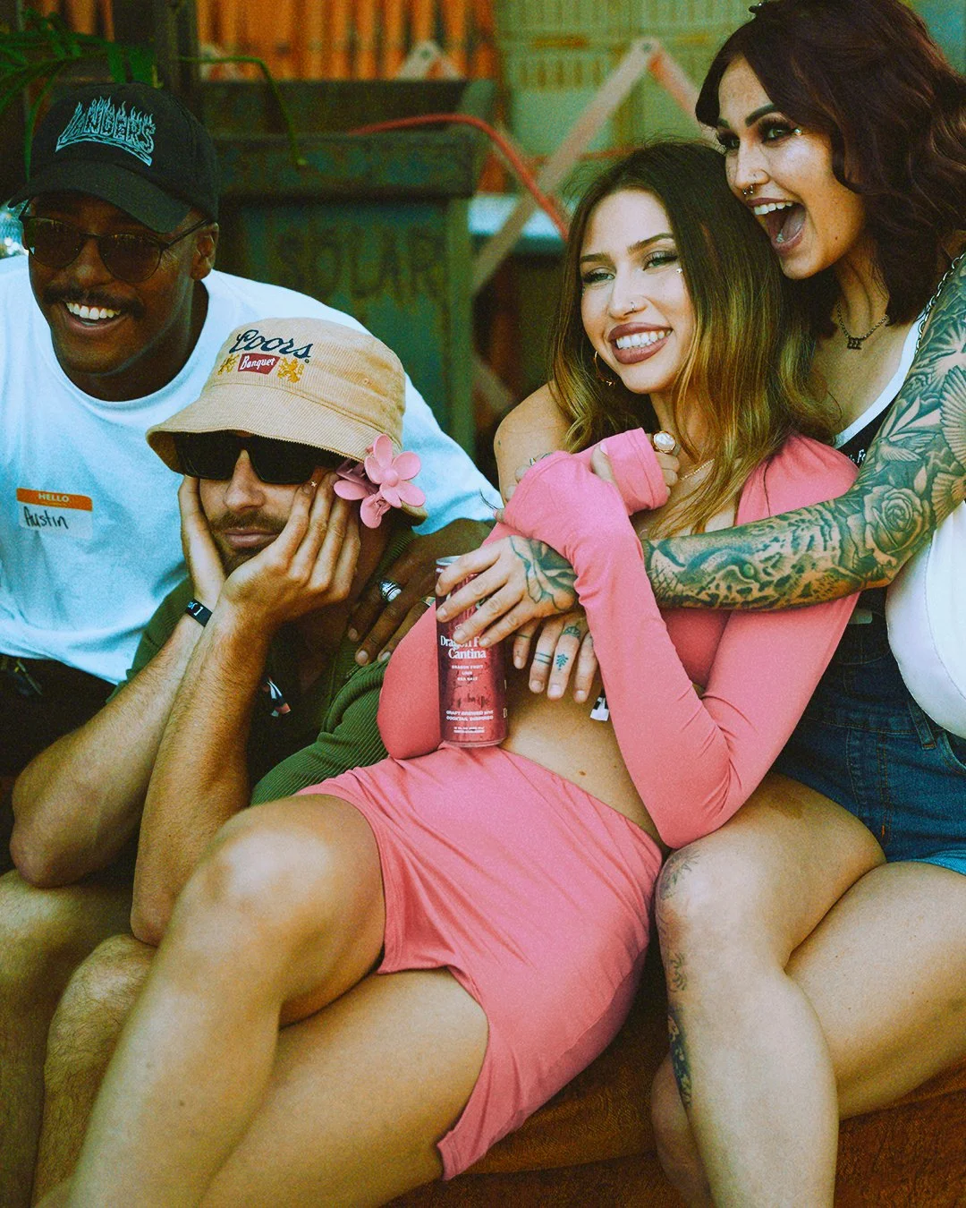 Group of five friends enjoying a relaxed moment together in a colorful outdoor setting. One woman in pink shorts and top with tattoos is sitting on a couch, holding a drink and smiling. The others are casually gathered around her, some with tattoos a