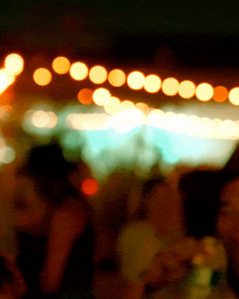 Blurry photograph of people at a nighttime event with string lights overhead and a large illuminated display in the background.