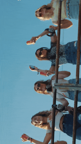 Five young women taking photos with smartphones on a boat with a dog also on the boat, against a clear blue sky.