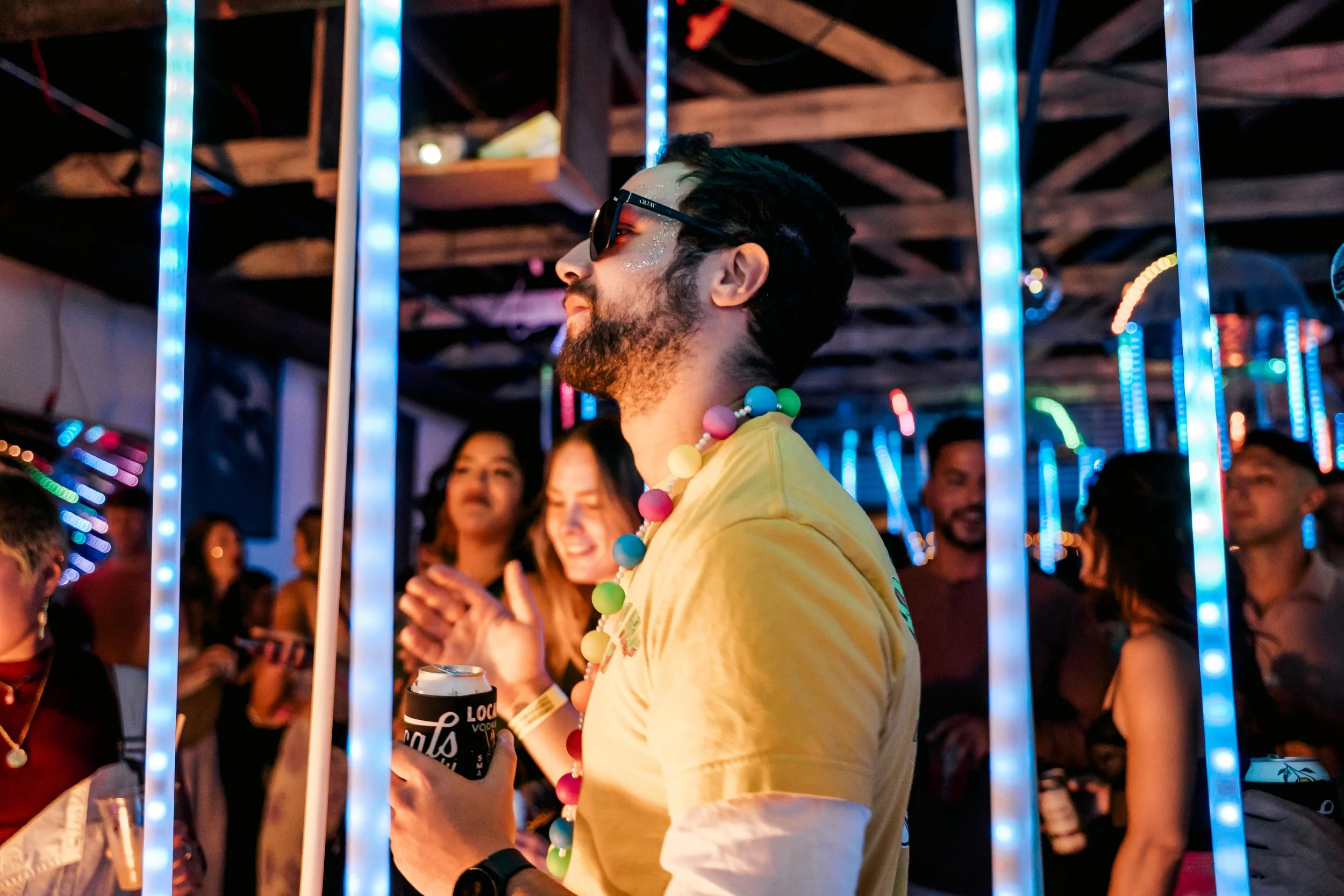 A man with dark hair, beard, and sunglasses, wearing a yellow shirt and a colorful bead necklace, holding a can, at a party with vibrant blue and pink neon lights and other people dancing and chatting in the background.