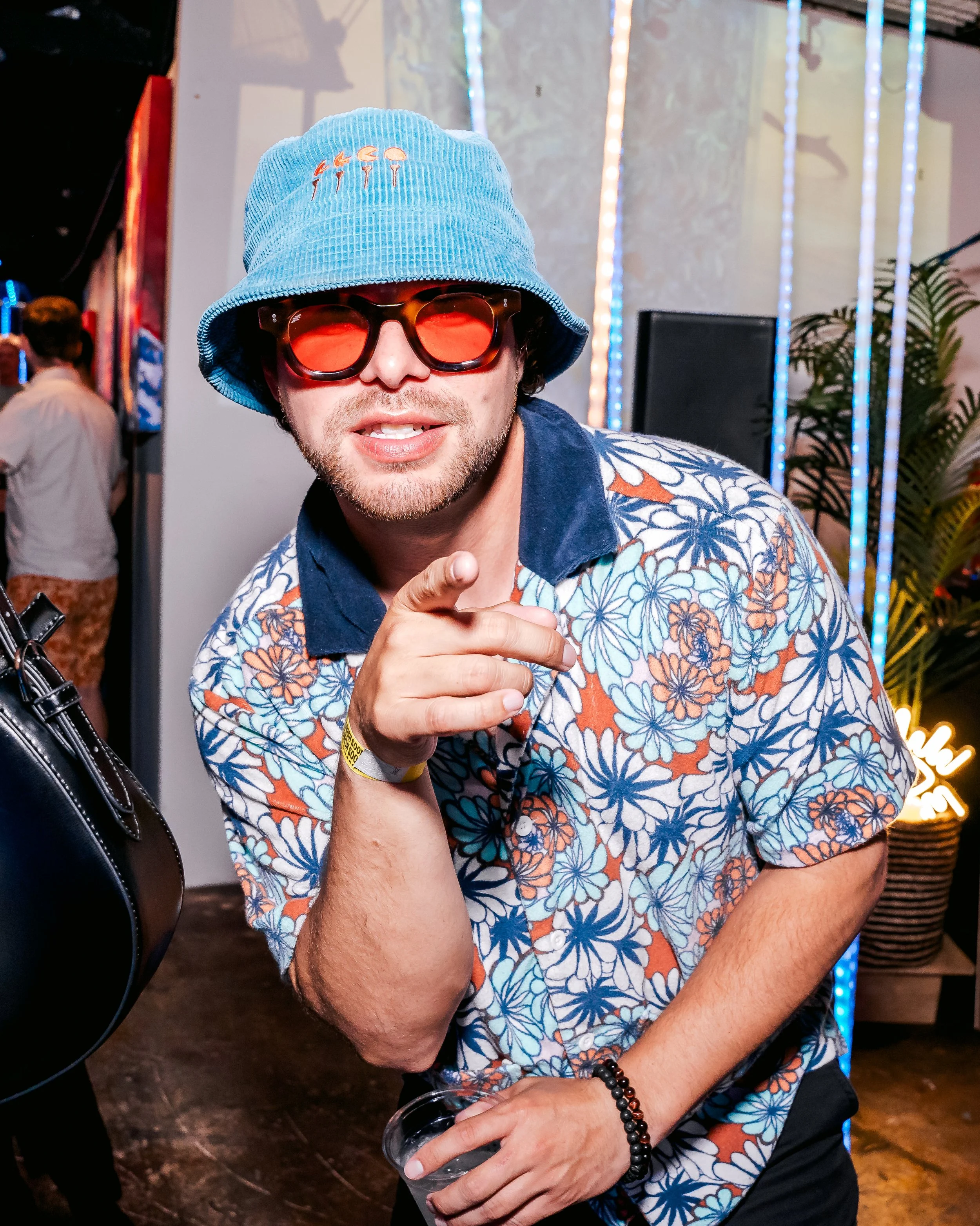 Man wearing a blue bucket hat, sunglasses with red lenses, and a colorful floral shirt at a party, pointing at the camera.