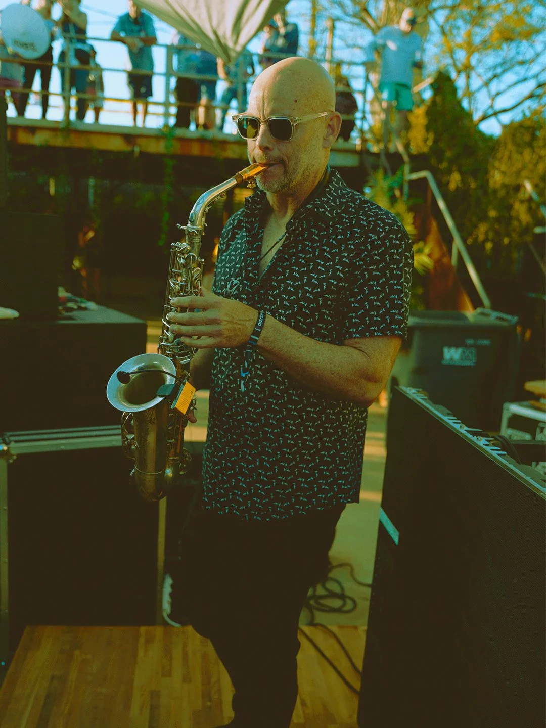 A bald man wearing sunglasses and a patterned shirt playing the saxophone outdoors, with people on a deck and trees in the background.