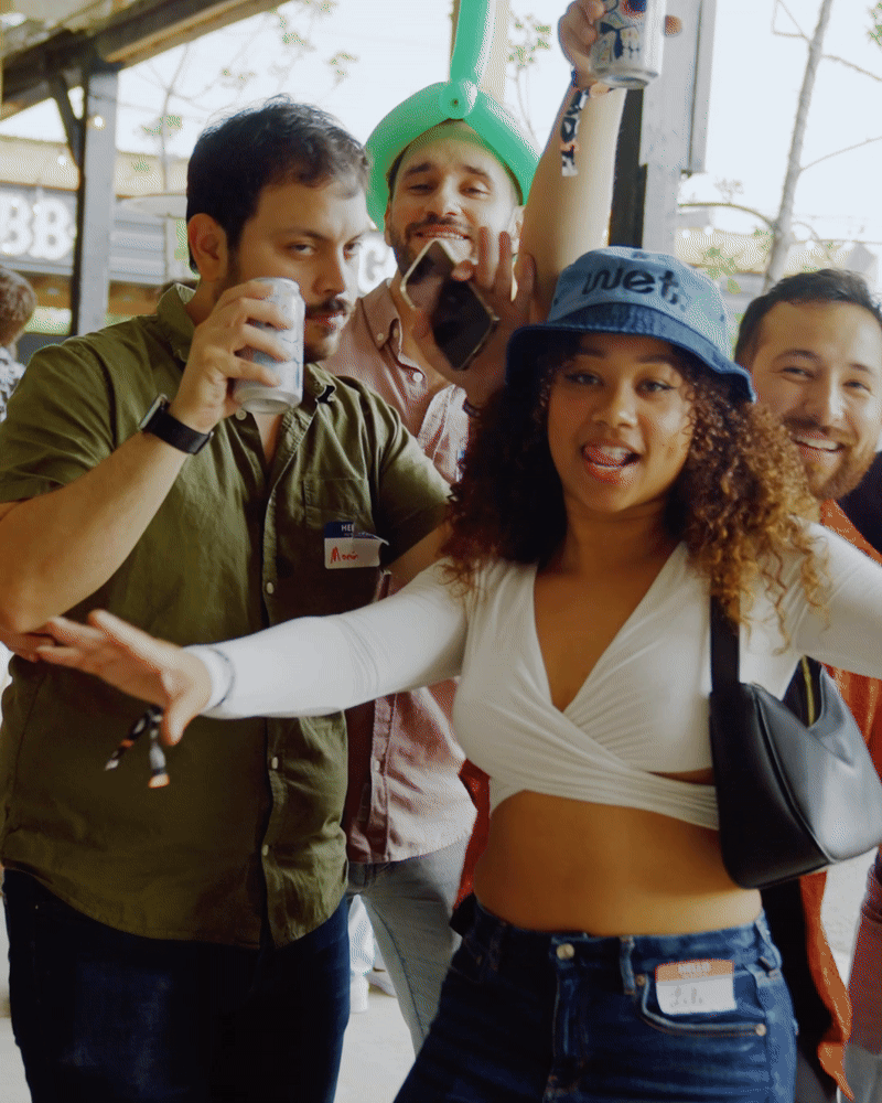 Group of young adults enjoying a party outdoors, some holding drinks, one wearing a green hat, and a woman dancing in the foreground.