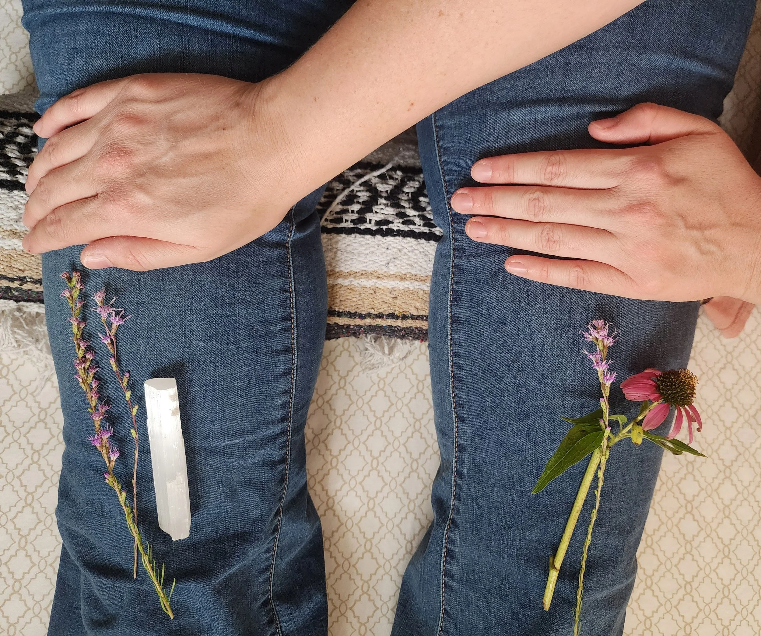 Reiki client is wearing blue jeans sitting on a patterned surface with multiple flowers on their lap and a white stick on the left side of the image.