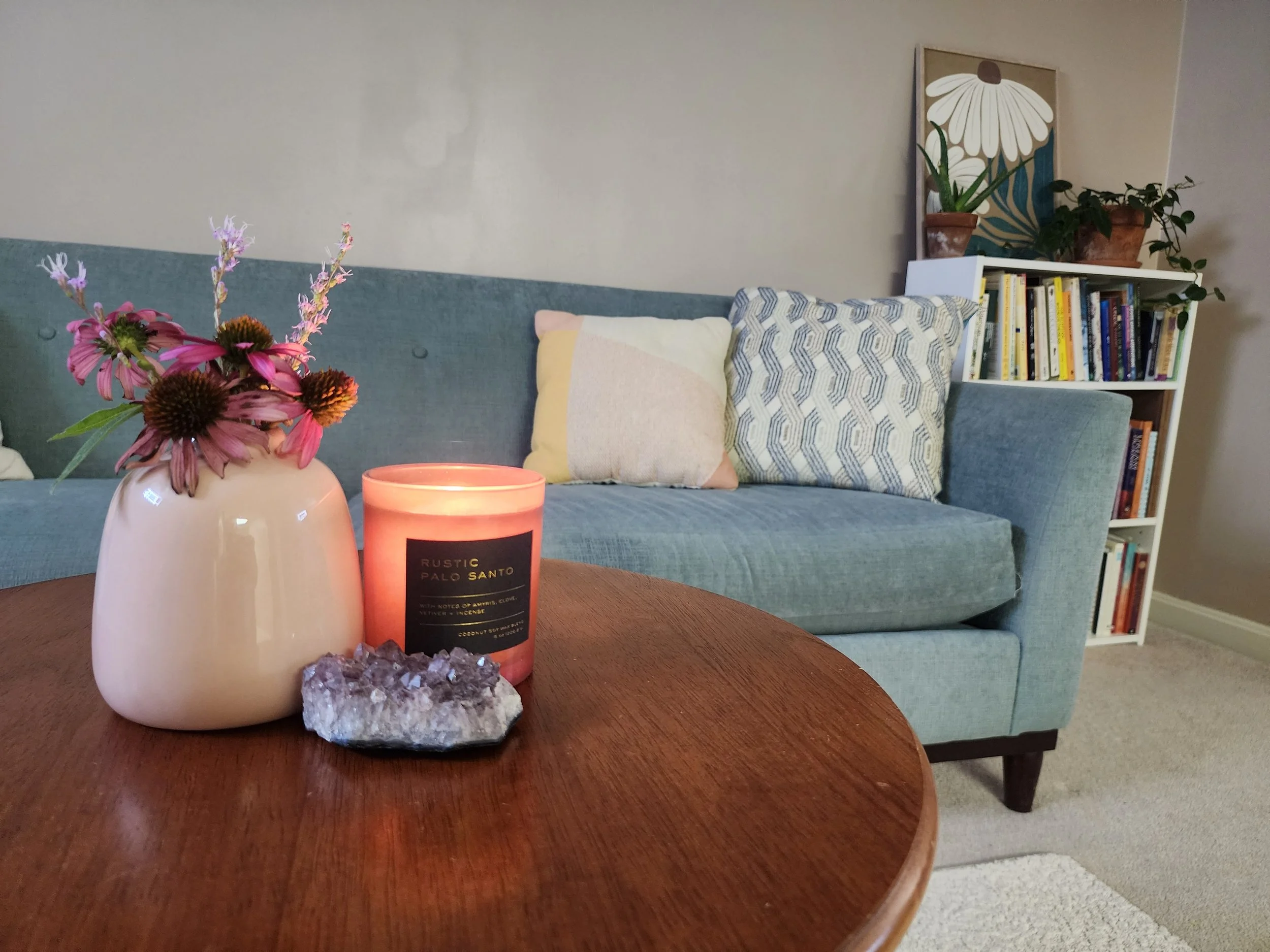 Close-up of a wooden table with a pink vase holding pink and purple flowers, a lit candle labeled 'Rustic Palo Santo,' and a cluster of rocks. In the background, there's a blue sofa with pillows and a bookshelf filled with books, a potted plant, and wall art featuring large white daisies.
