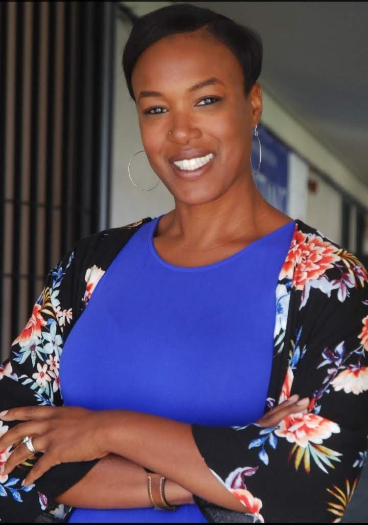 Joanna Paun wearing a blue top and a black floral shawl, standing with crossed arms in an indoor setting.