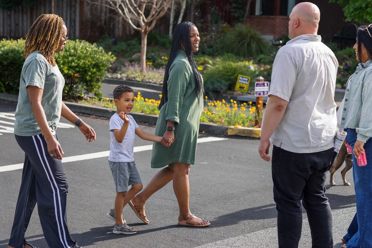 Group of five people crossing a street, including a woman holding a child's hand, smiling and talking, with a woman holding a water bottle in the background.