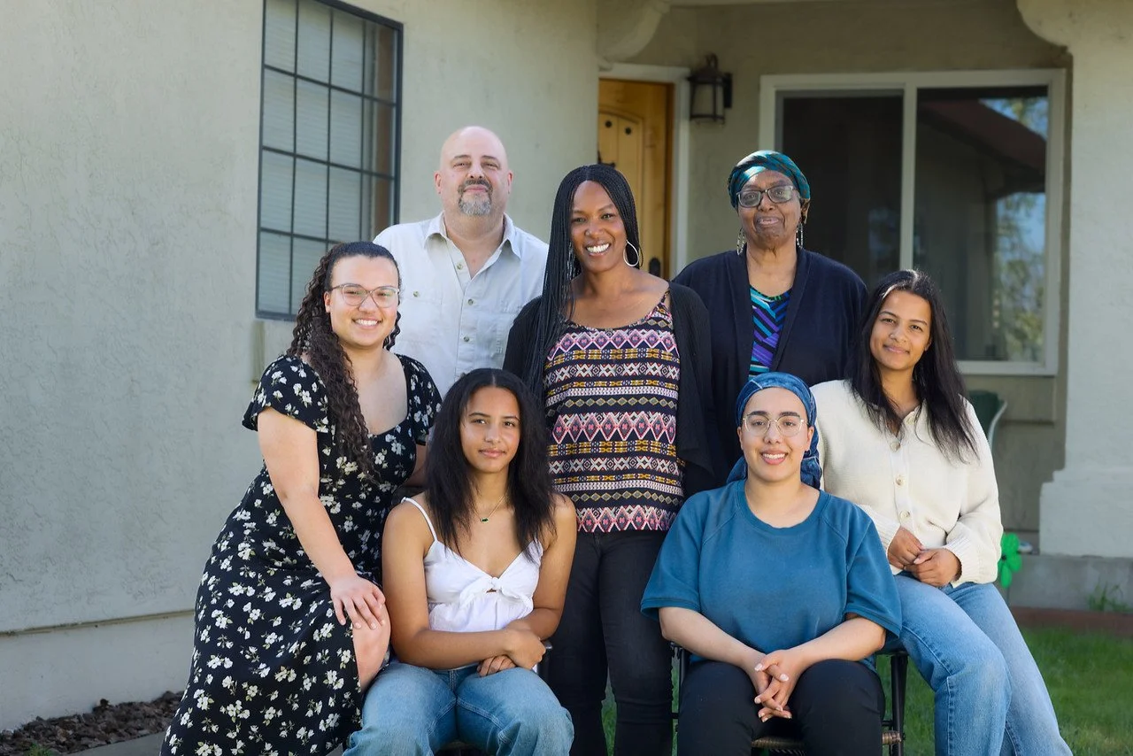 Group of eight diverse people standing and sitting outside a house, smiling.