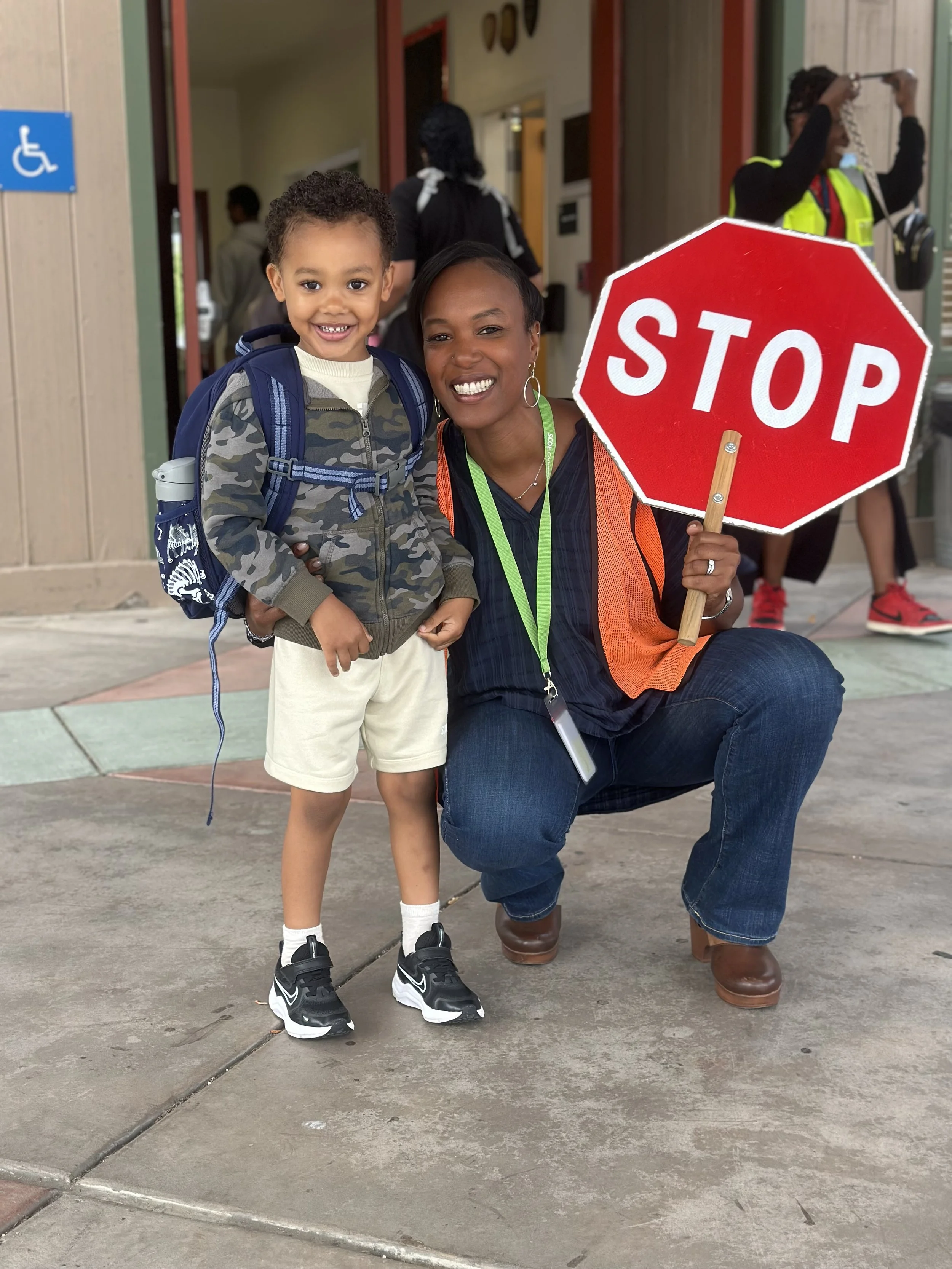A young boy smiling and standing with Joanna Paun, who is kneeling and holding a stop sign. The boy has a backpack and is dressed in casual clothes. The woman is wearing a safety vest and has a lanyard with a badge.