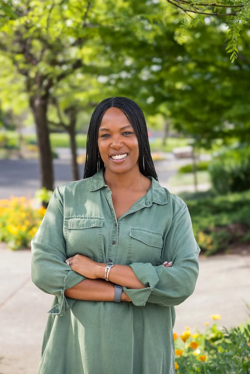 A woman with braided hair smiling outdoors on a sunny day, wearing a green button-up shirt with her arms crossed, standing in front of trees and flowering bushes.