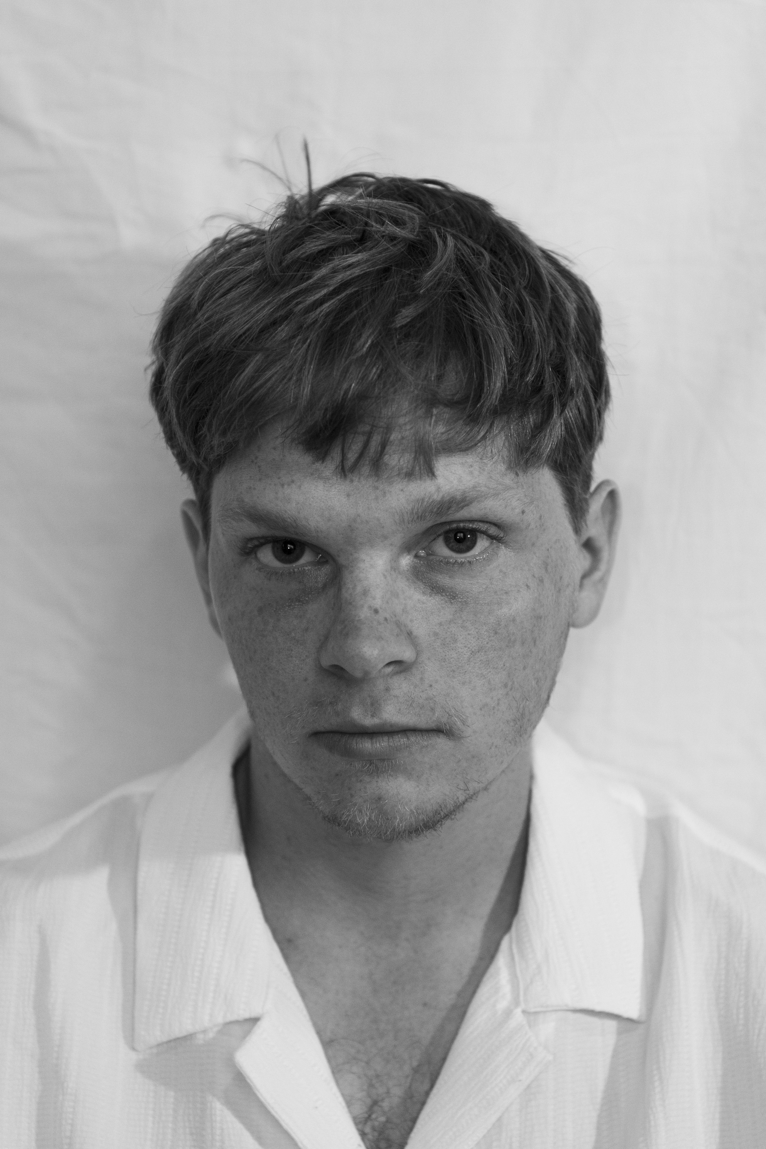 Black and white close-up photo of a person with short, messy hair and light skin, wearing a white shirt with a collar.