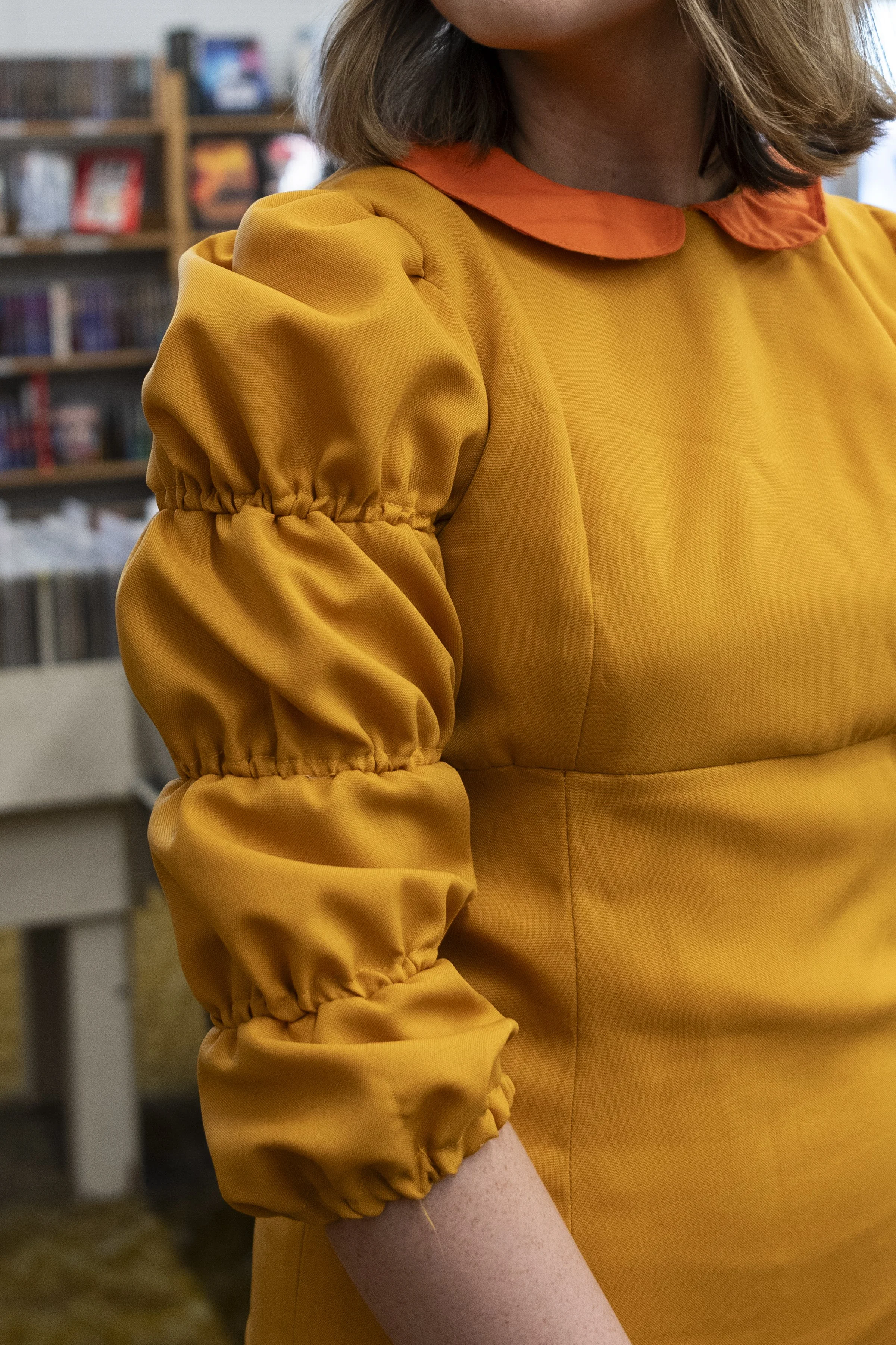 Close-up of a woman wearing a mustard yellow dress with puffed sleeves and an orange collar, standing in a room with bookshelves in the background.