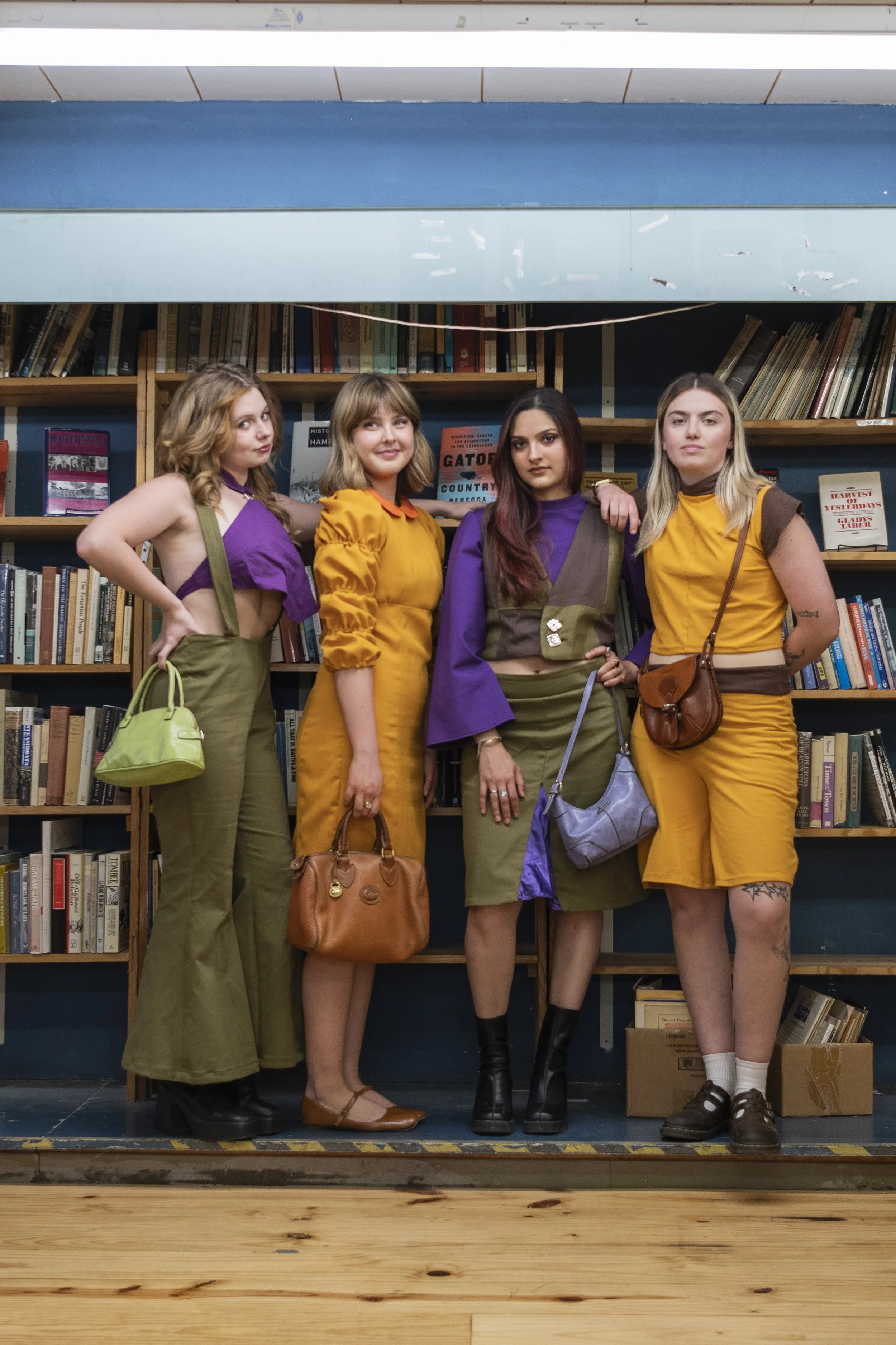 Four young women standing close together in front of a bookshelf, posing for a photo. They are dressed in colorful, vintage-style outfits, carrying various handbags, and standing on a small stage in a room with wooden floors.