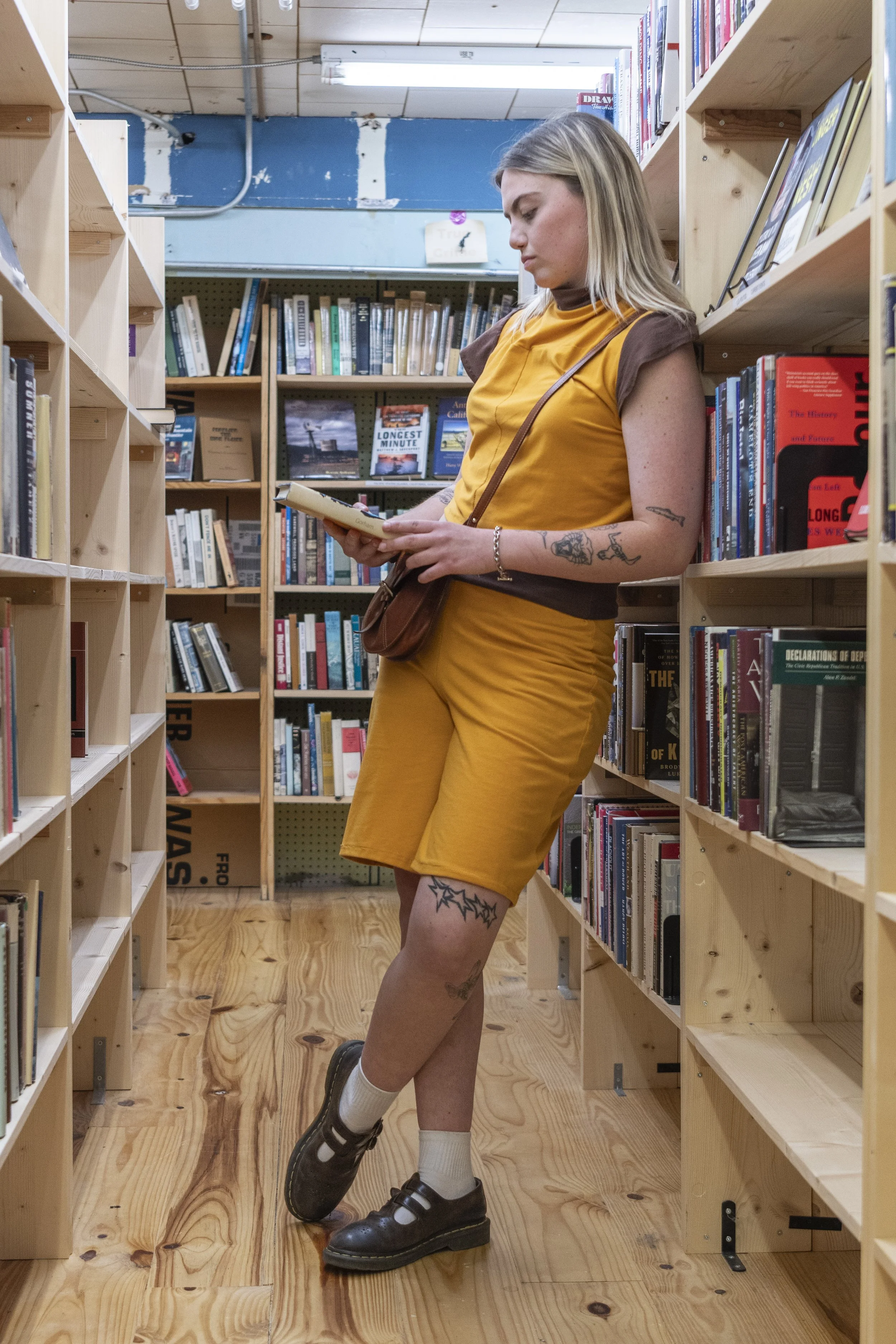 A young woman with tattoos on her arm and leg, wearing a yellow dress, white socks, and black shoes, stands in a bookstore aisle reading a book. She has blonde hair and is carrying a brown purse, with bookshelves filled with books on both sides of her.