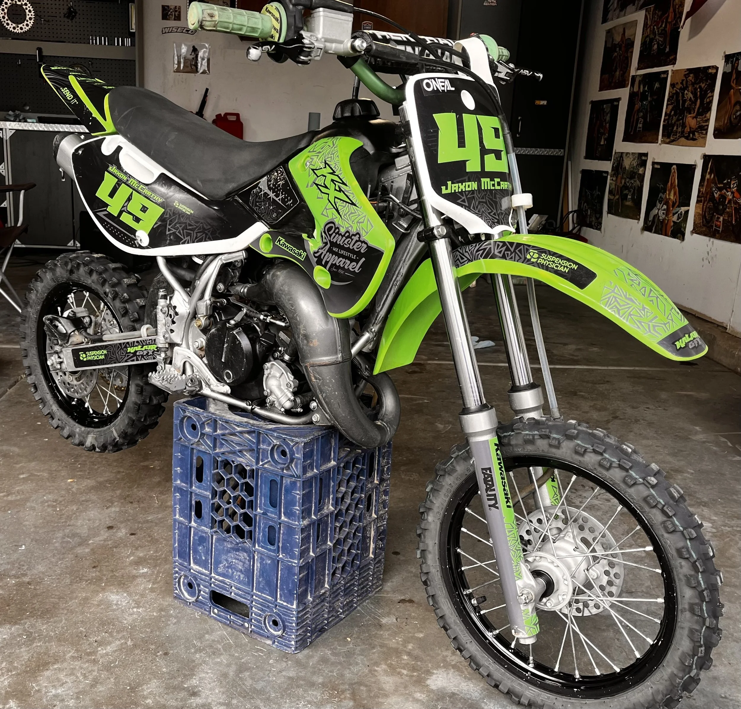 A green and black Kawasaki dirt bike with racing number 49, sitting on a blue crate in a garage. The bike has decals and branding, including suspension, physique, and apparel stickers, with posters on the garage walls.