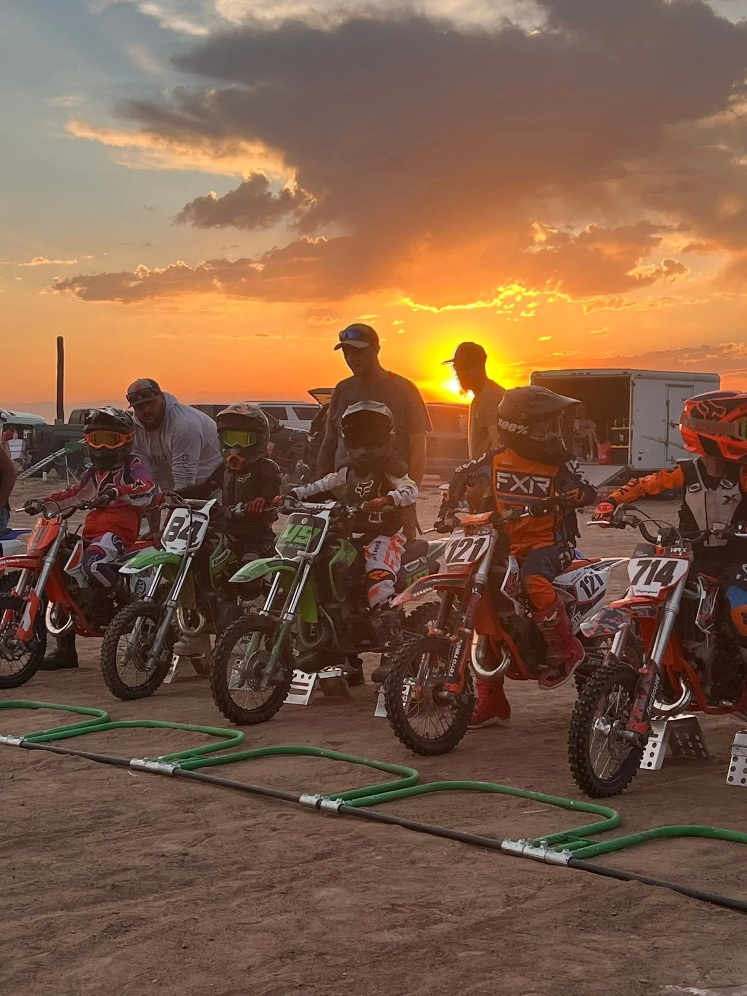 Kids and adults on dirt bikes at sunset, preparing for a motocross race, with green hoses on the dirt ground.