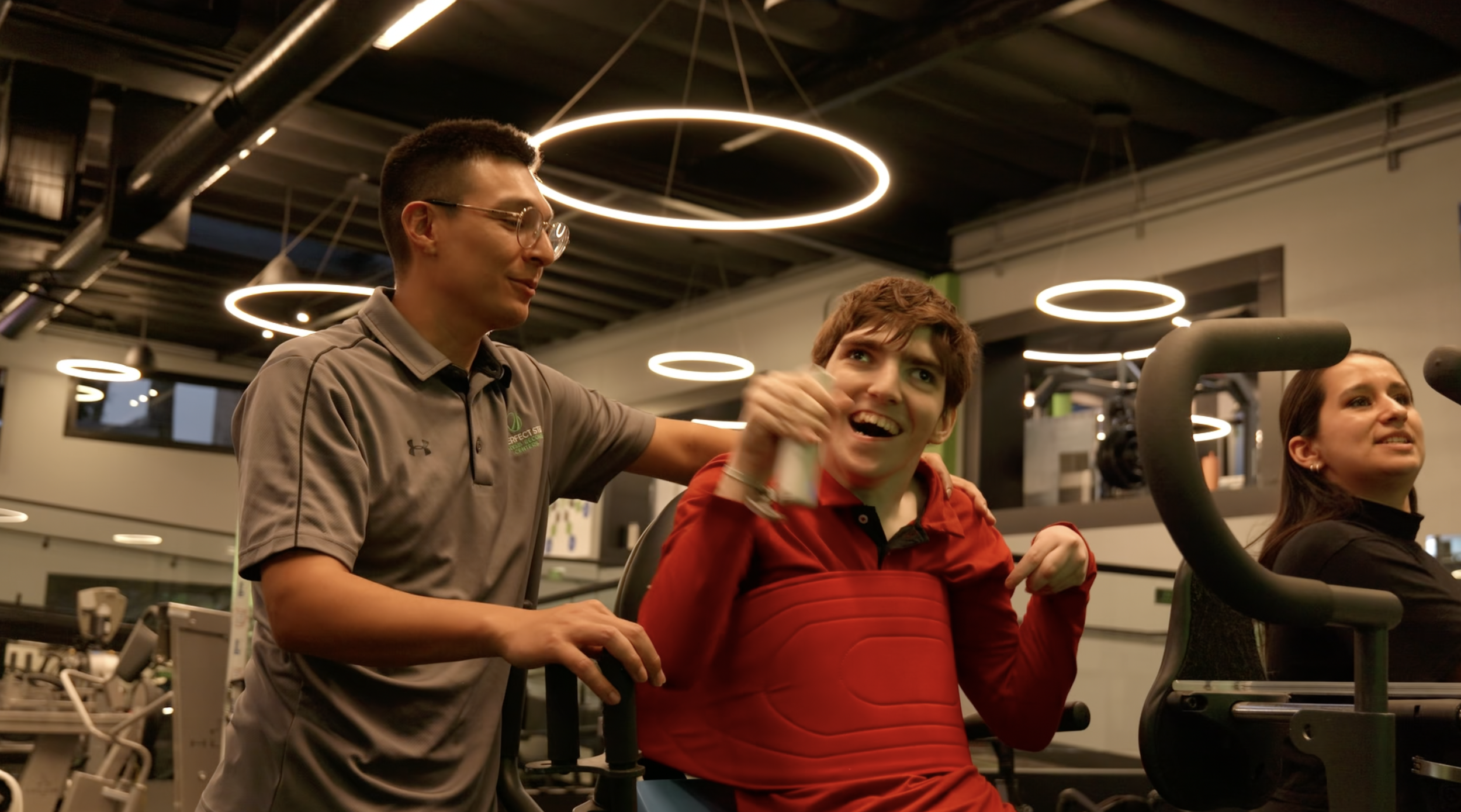 A young man with short brown hair and a red shirt is celebrating at a gym with a staff member in a gray polo shirt. Film production still from brand film directed by Jhovany Quiroz Studio