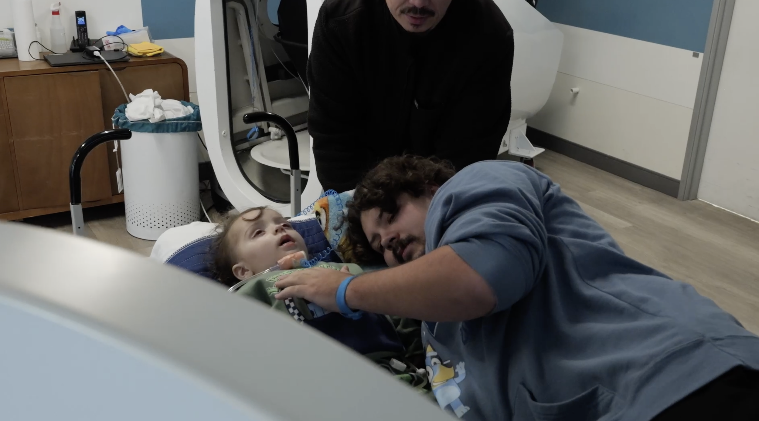 A young girl lying in a hospital bed with medical tubes, conveying as a caring man leans over her, and another man stands nearby. Film production still from brand film directed by Jhovany Quiroz Studio