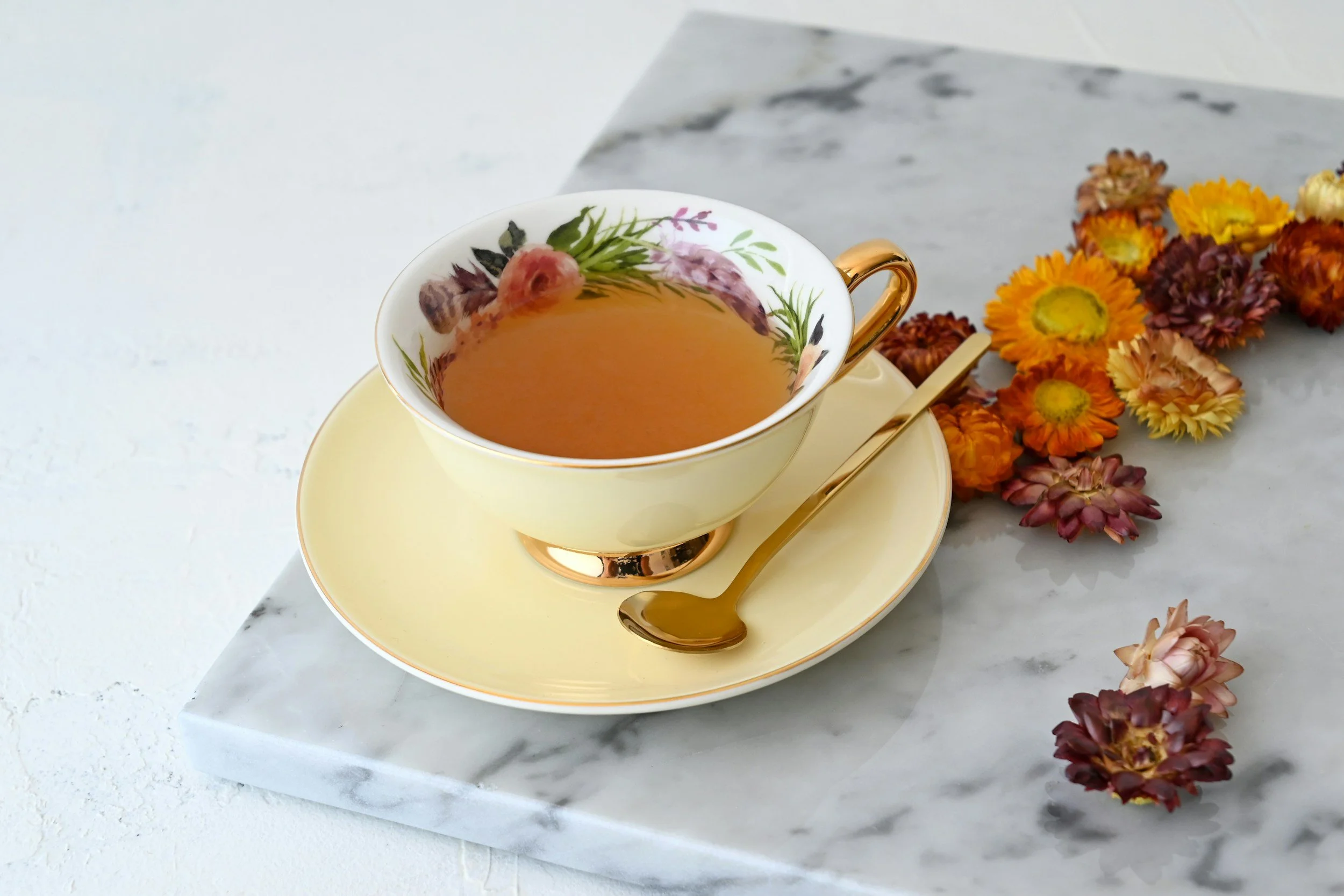 A teacup with floral design and gold trim filled with tea, placed on a matching saucer with a gold spoon, on a marble slab, with dried colorful flowers beside it.
