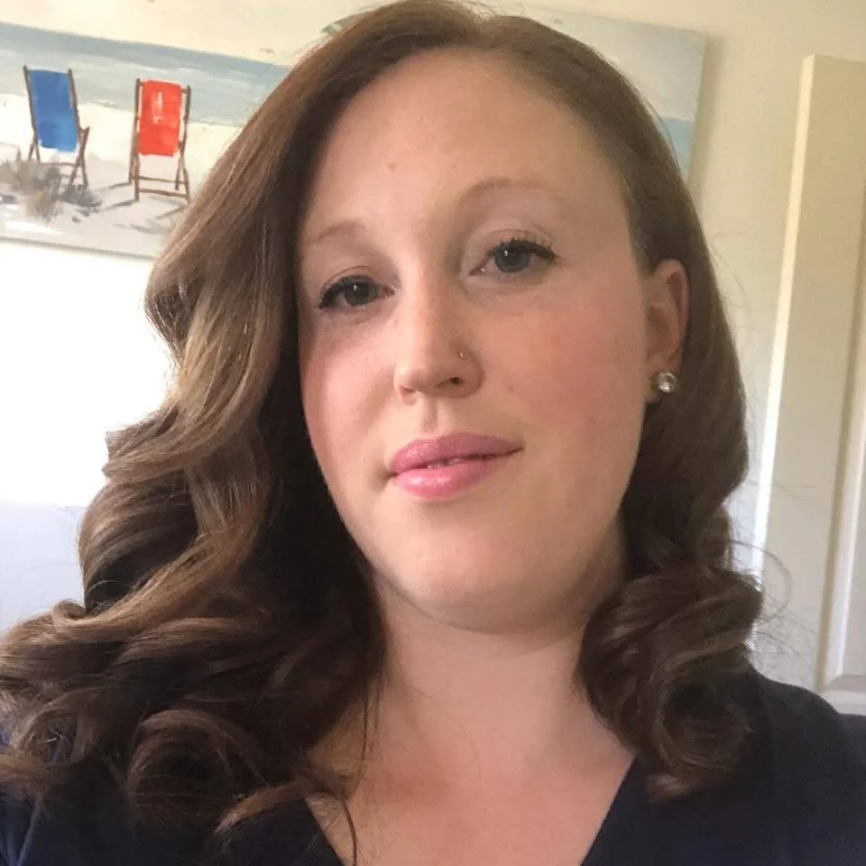 A woman with wavy brown hair, light skin, and a nose piercing taking a selfie indoors.
