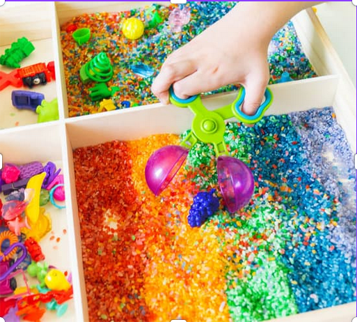Child playing with a colorful rainbow-themed sensory bin filled with beads and small toys, using a green hand-held scoop with purple bowls.