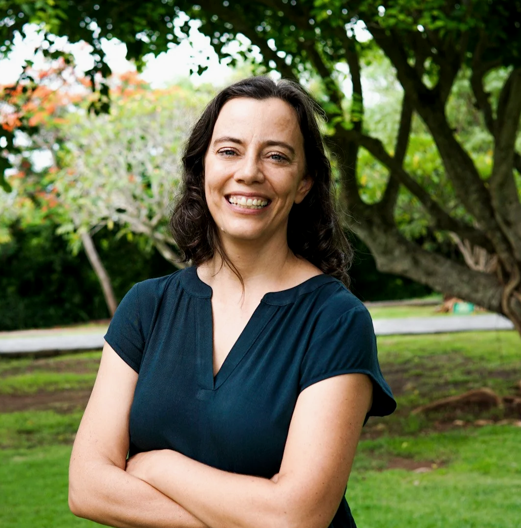 A woman with dark brown hair smiles with arms crossed outdoors, standing in front of a large tree and greenery.