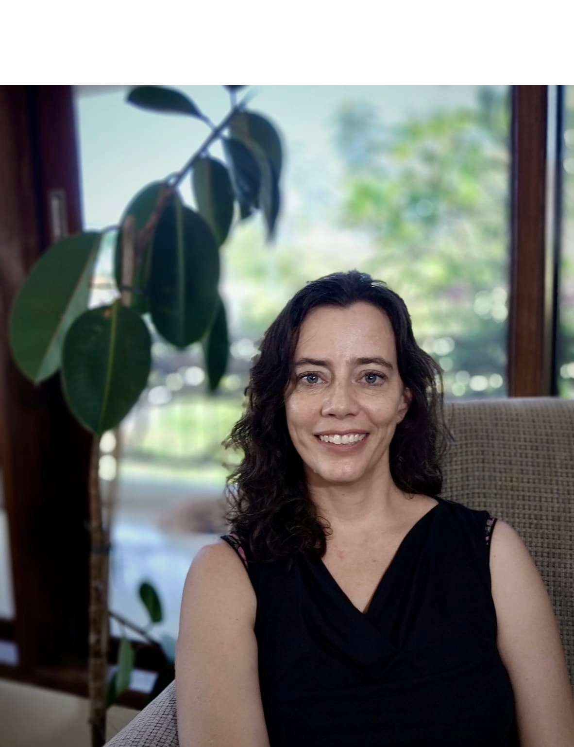 Smiling woman sitting in a living room near a large window with greenery outside, and a tall potted plant in the background.
