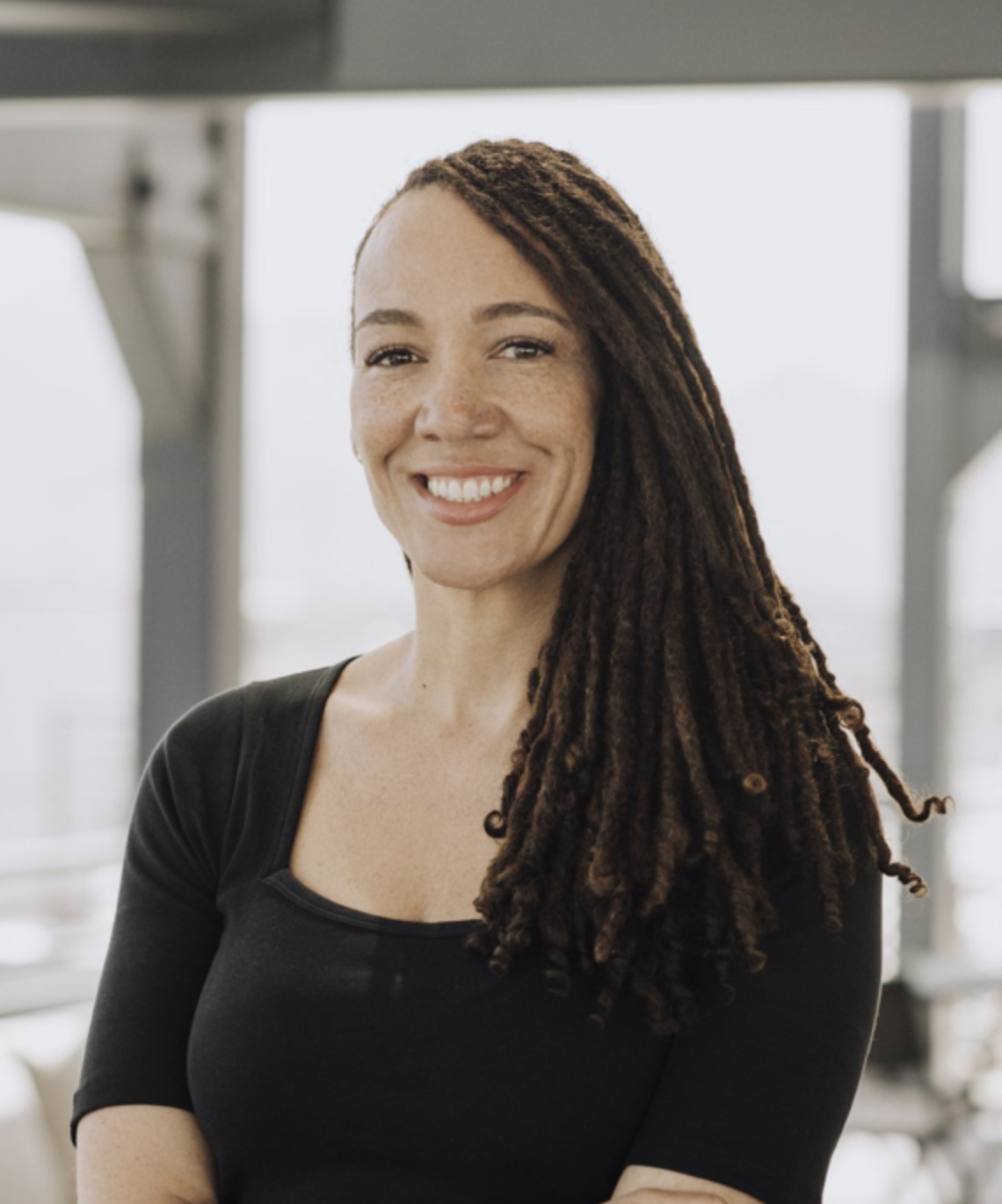 A woman with long, styled dreadlocks smiling in an indoor setting with large windows.
