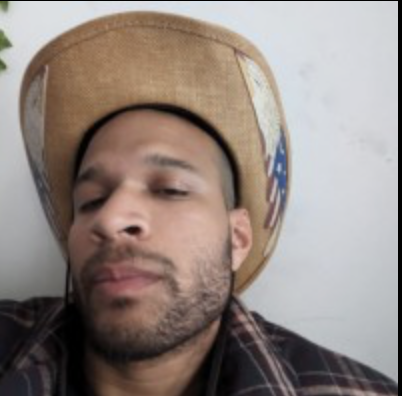 Close-up of a man wearing a cowboy hat, with a plaid shirt, looking slightly downward.