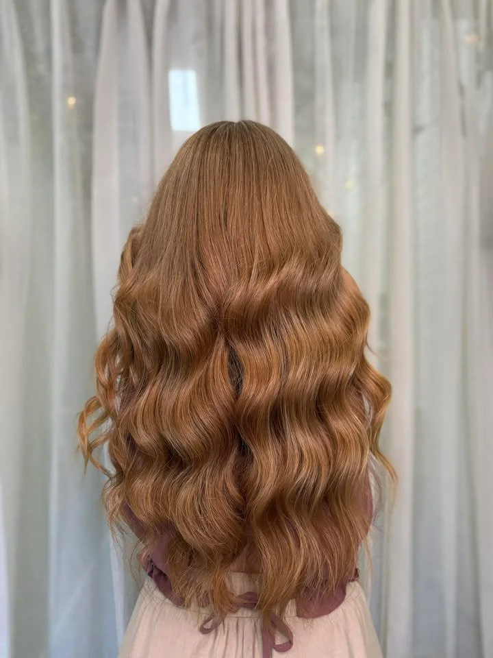 Back view of a woman with long, wavy, auburn hair standing in front of light-colored curtains.