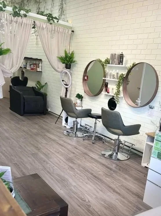 Interior view of a hair salon with two styling chairs and mirrors on a white brick wall, featuring shelves with hair products, potted plants, and a black armchair in the corner, with light wood flooring and sheer curtains in the background.