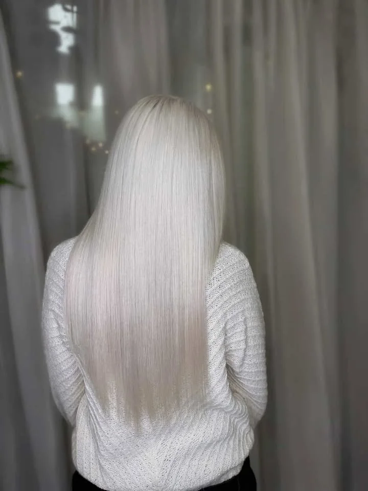 Person with long, straight, platinum blonde hair, wearing a white knit sweater, standing indoors with curtains in the background.