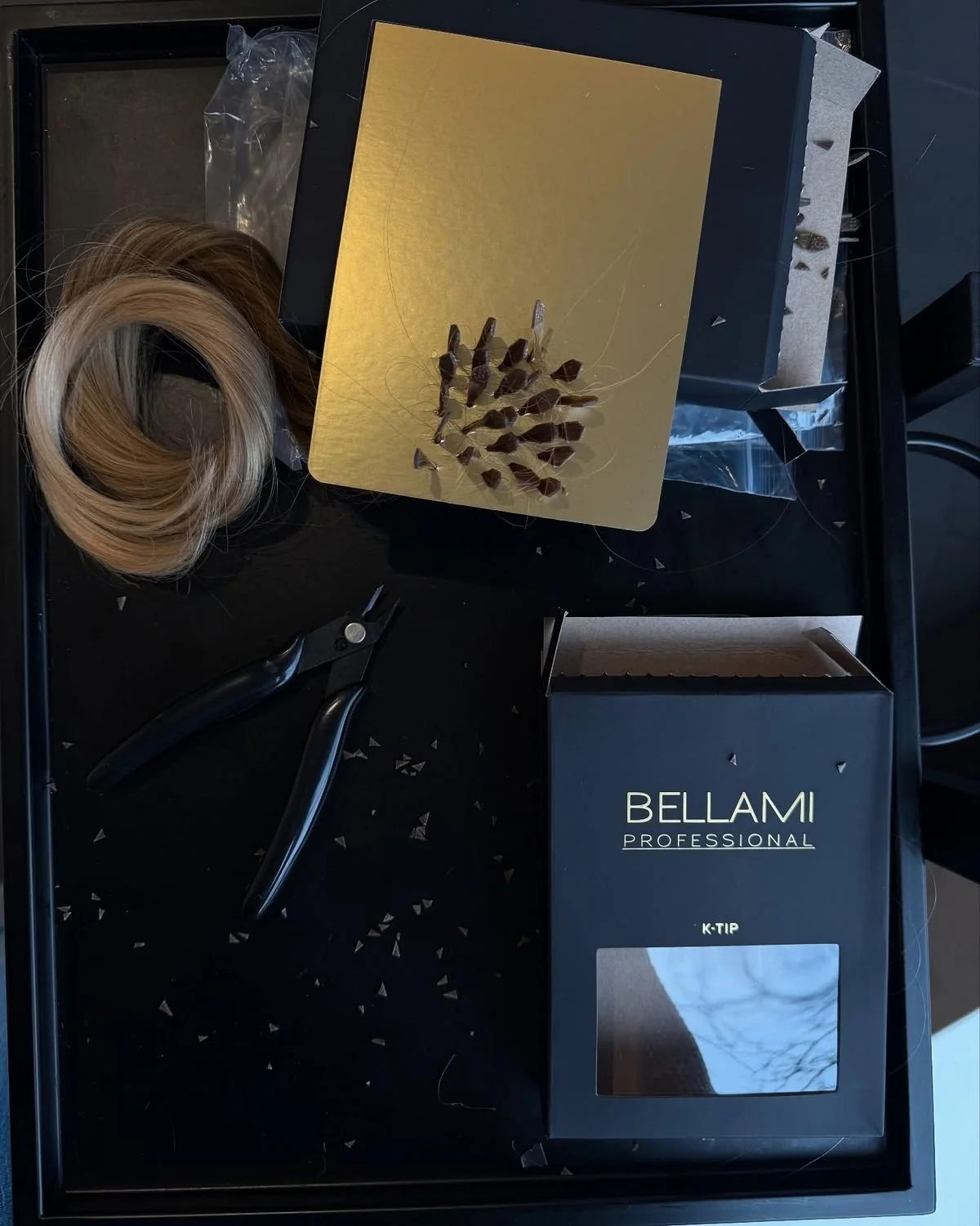 Hair extensions, a pair of pliers, a black and gold box, and a gold-colored mat with hair extensions attached, arranged on a black table.