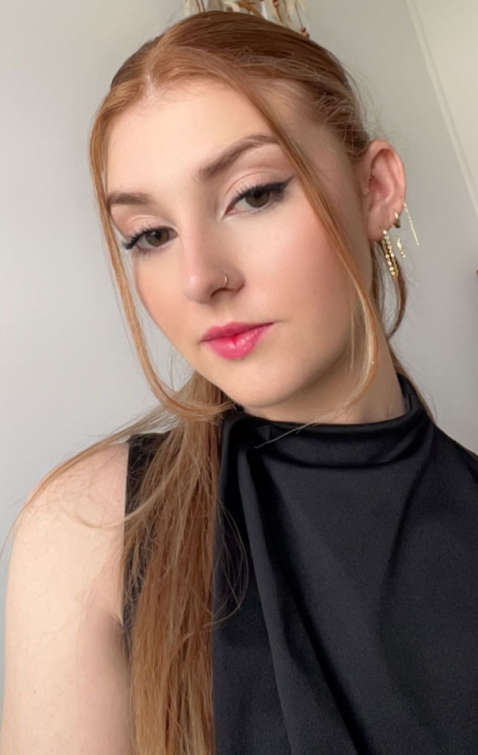 A young woman with red hair and fair skin looking into the camera, wearing a black sleeveless top with a high neckline, gold earrings, and a nose piercing, in front of a plain light-colored wall.