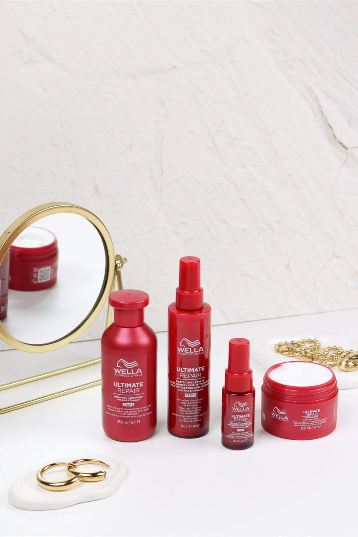 Red hair care products labeled Wella Ultimate Repair, including shampoo, leave-in spray, and treatment cream, arranged on a white surface with a gold mirror, jewelry, and a white ceramic dish.
