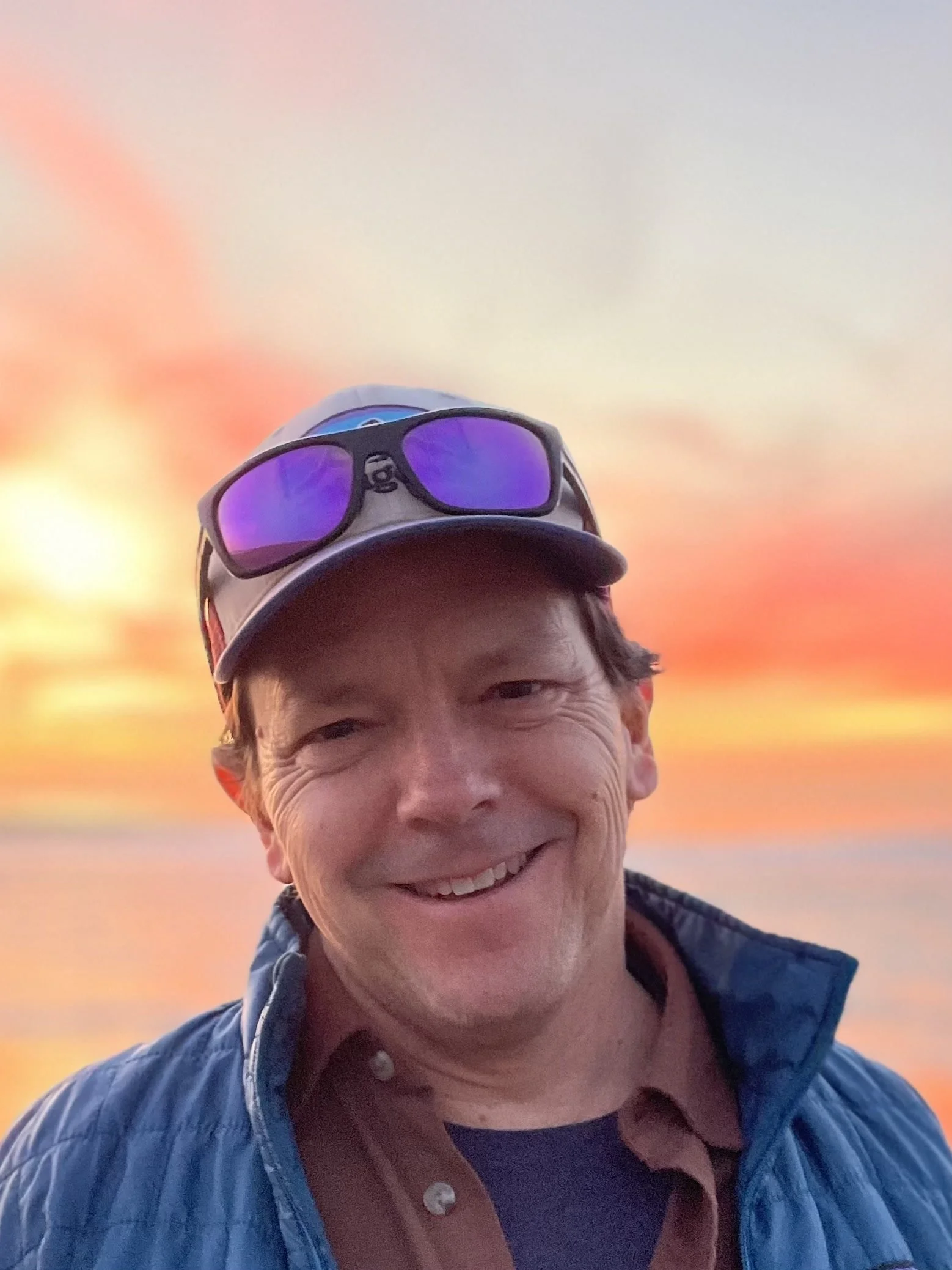 A smiling man wearing sunglasses and a baseball cap, outdoors at sunset with colorful sky in the background.