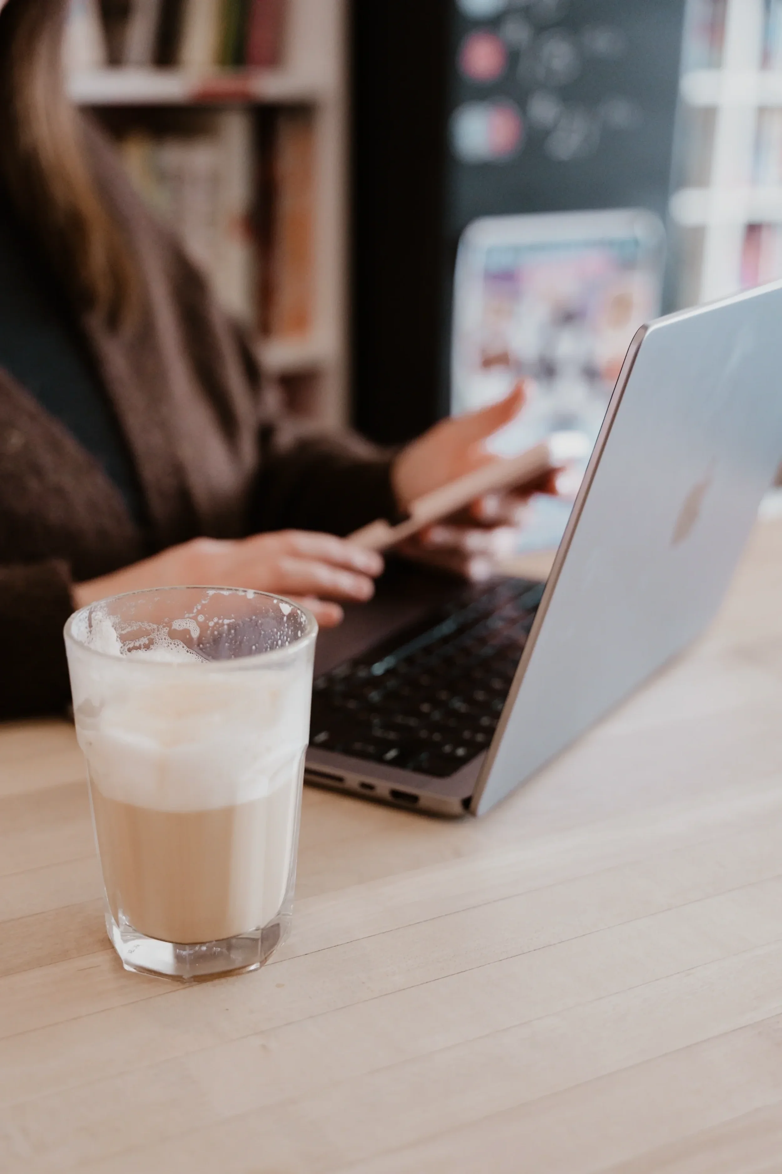 A glass of iced coffee on a wooden table in front of a person using a laptop and a tablet in a cozy cafe.