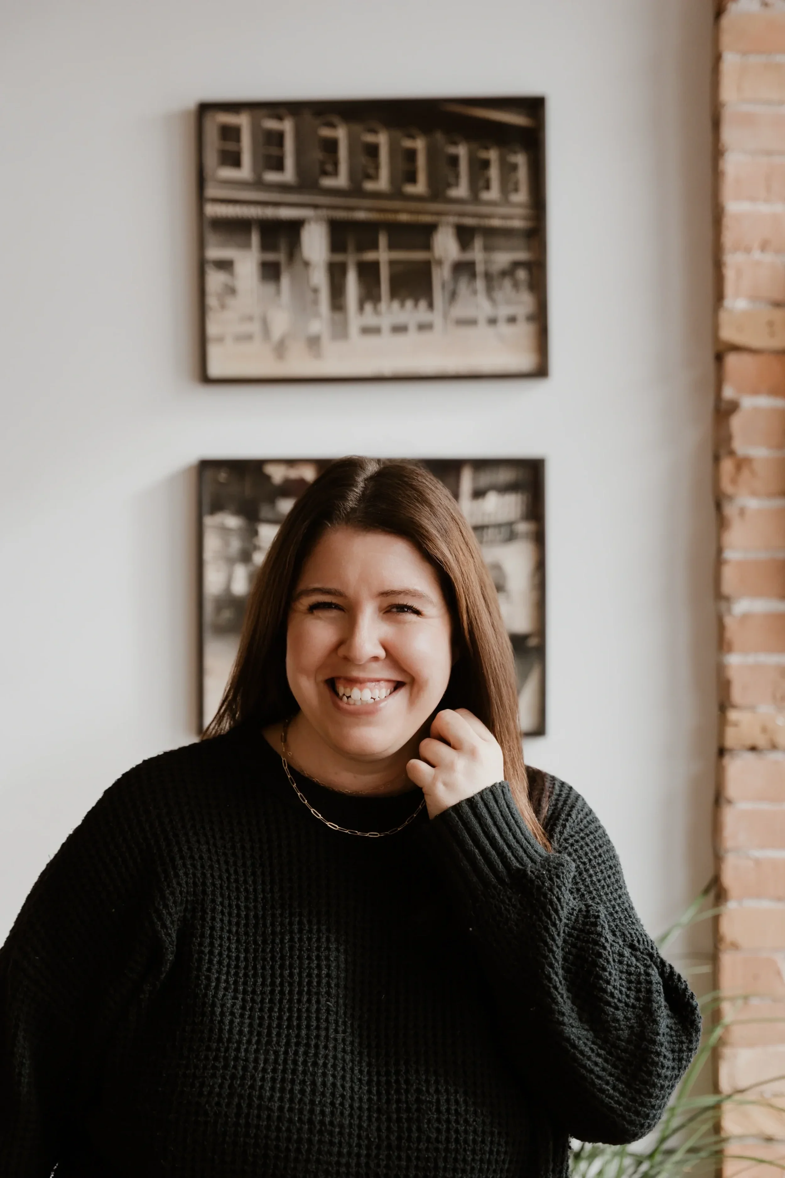 Amanda of Her Truth standing in front of a white wall with framed photographs, and a brick wall to her right.