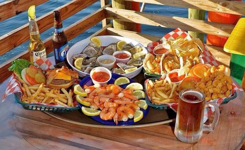 Seafood platter with shrimp, oysters, with lemon wedges, fries, a burger with lettuce and tomato, fried fish, and two bottles of beer on a wooden table at an outdoor setting.