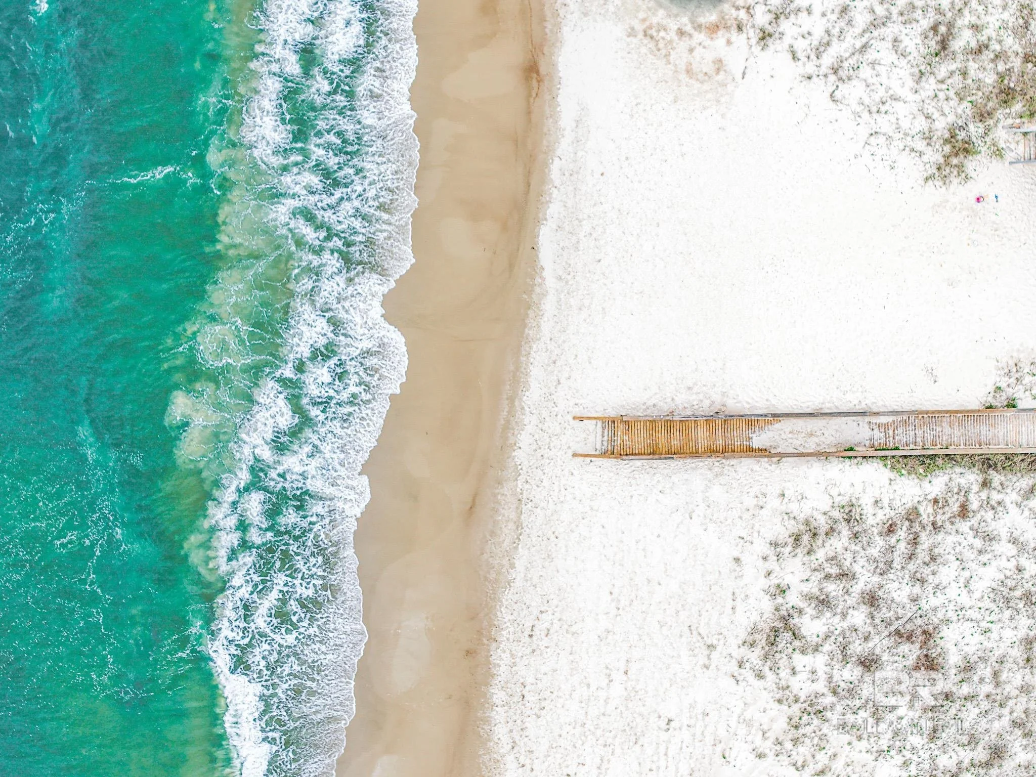 Aerial view of a sandy beach with turquoise waves washing ashore and a wooden walkway leading to the beach, with some trees and bushes on the right side.