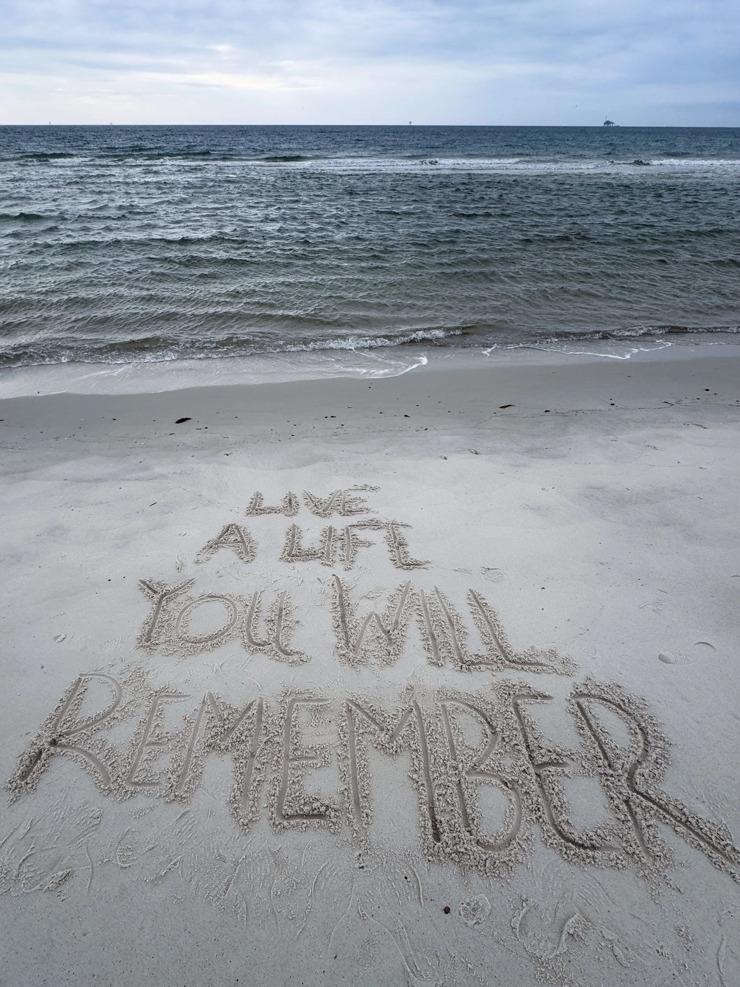 Message written in the sand on a beach that says, 'LOVE A LIFE YOU WILL REMEMBER,' with the ocean and cloudy sky in the background.