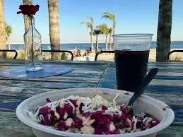A bowl of food on a table at a beachside setting with palm trees, a glass of soda, and a bottle of water in the background