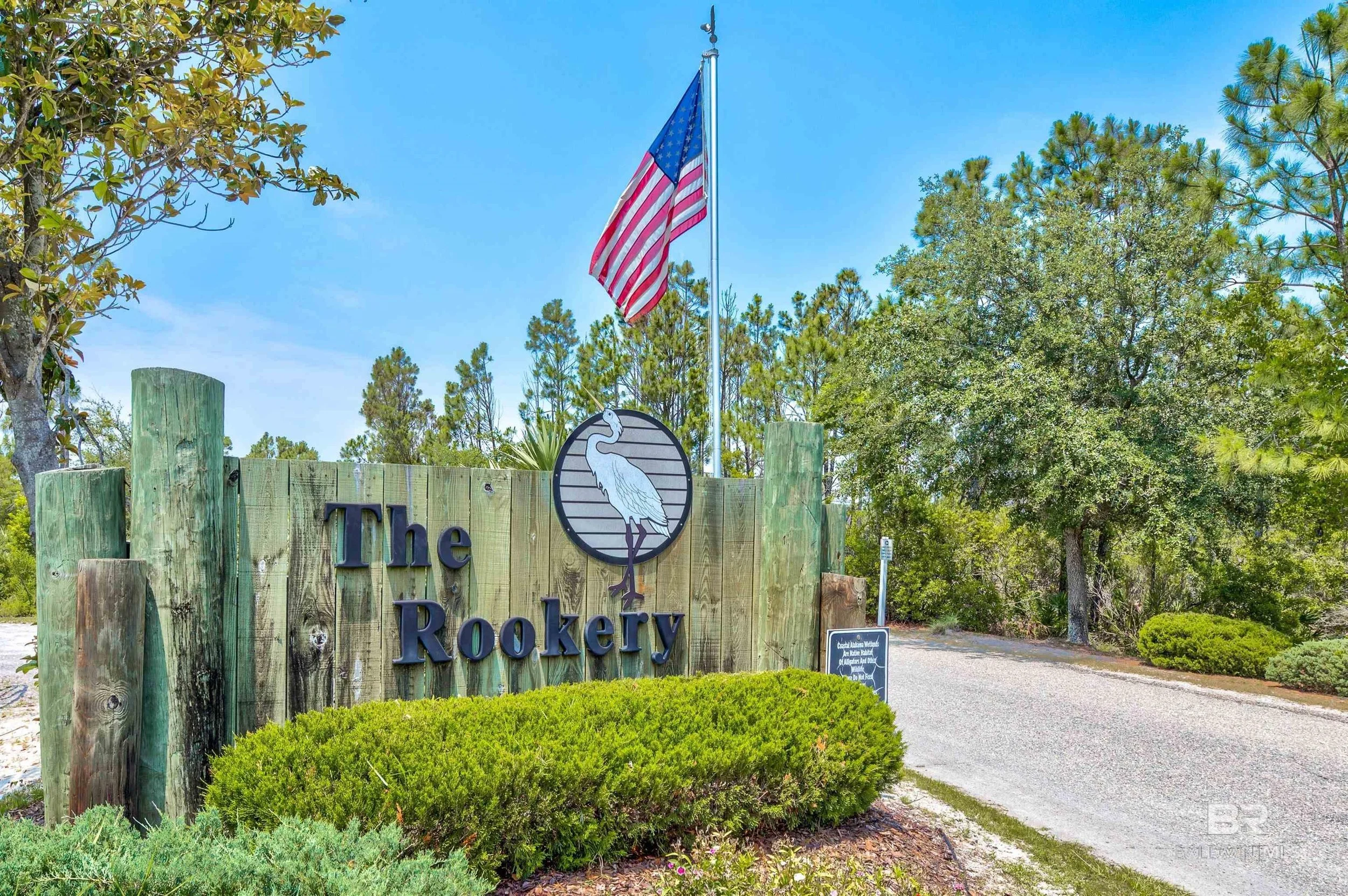 Entrance sign for The Rookery, featuring a wooden sign with a heron logo, an American flag flying, and trees in the background under a blue sky.