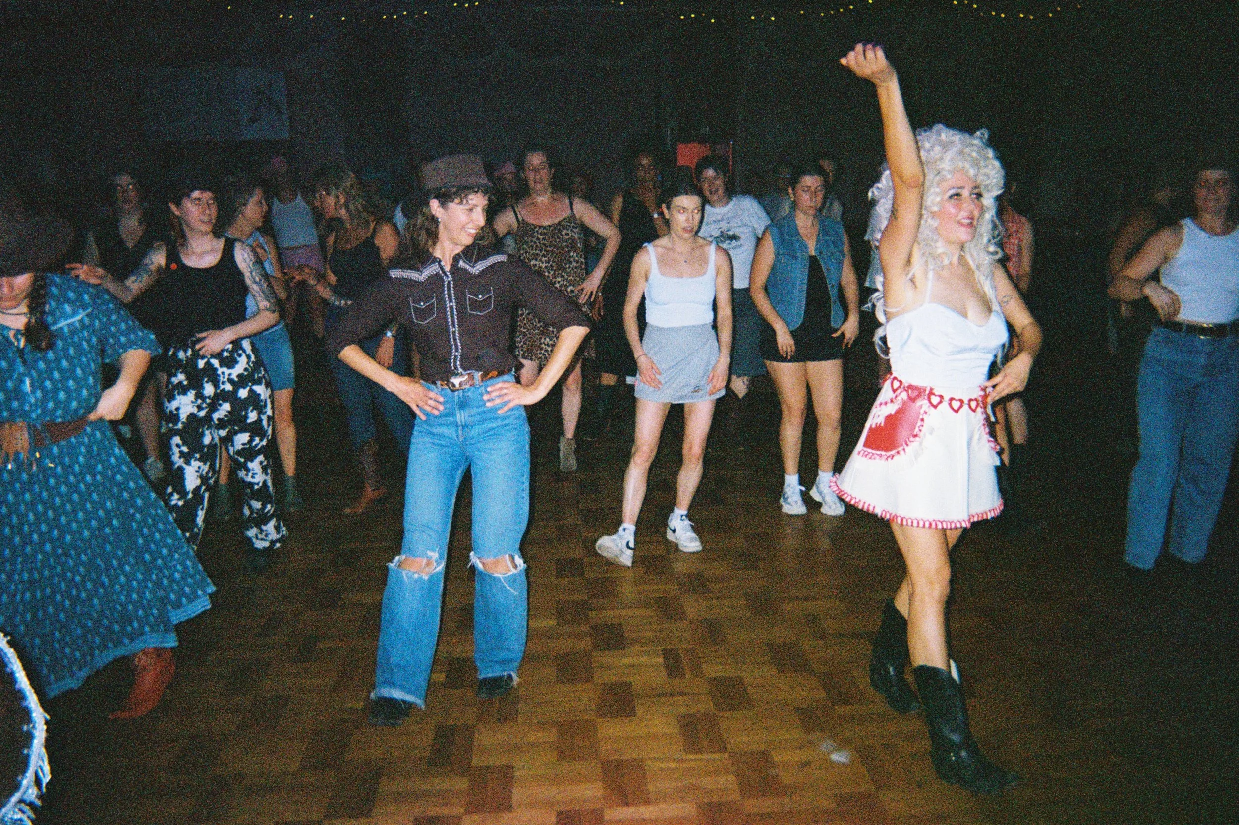 A group of women dancing at a party or dance event, with one woman dressed in a white outfit with heart decorations and a wig, leading the dance with her right arm raised.