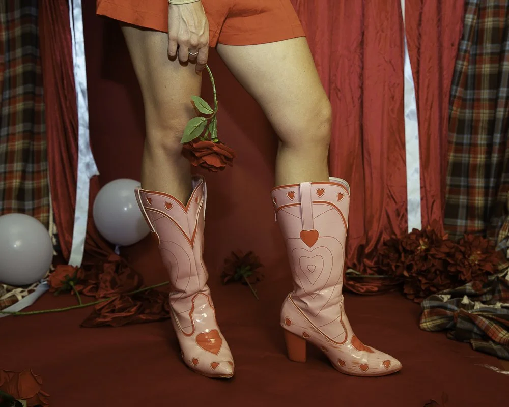 Person standing with legs apart, holding a red rose in one hand, wearing knee-high pink boots with heart designs, surrounded by red roses, white balloons, and red and plaid curtains.